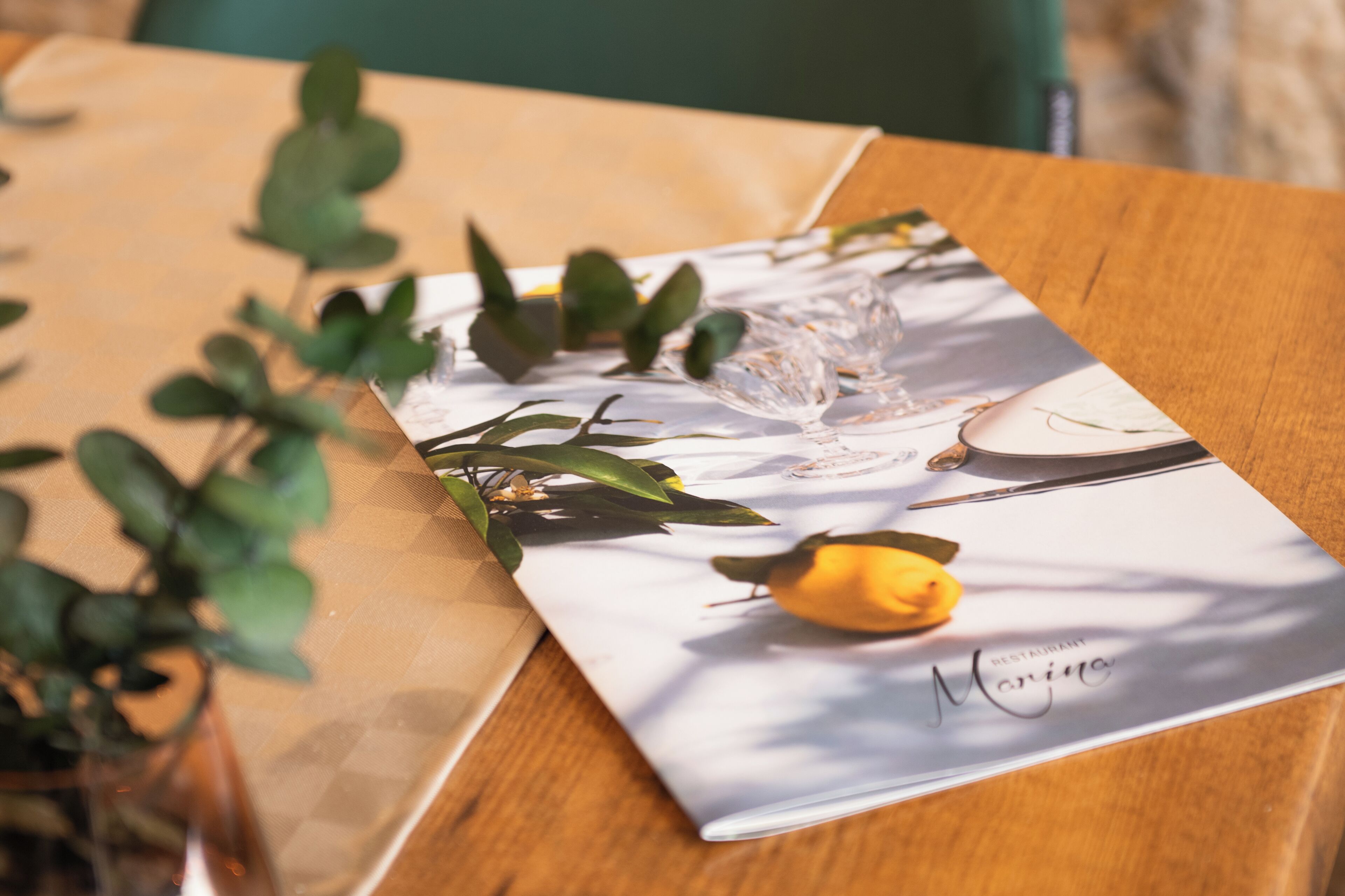 Une table avec un set de table orné du nom d’un restaurant, un vase en verre et des feuilles vertes.