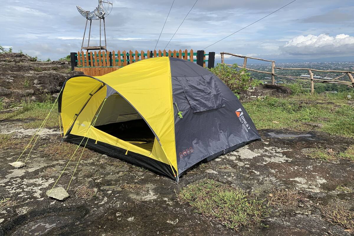 A yellow and gray tent set up on a rocky terrain with a wooden fence and a view of a distant cityscape.