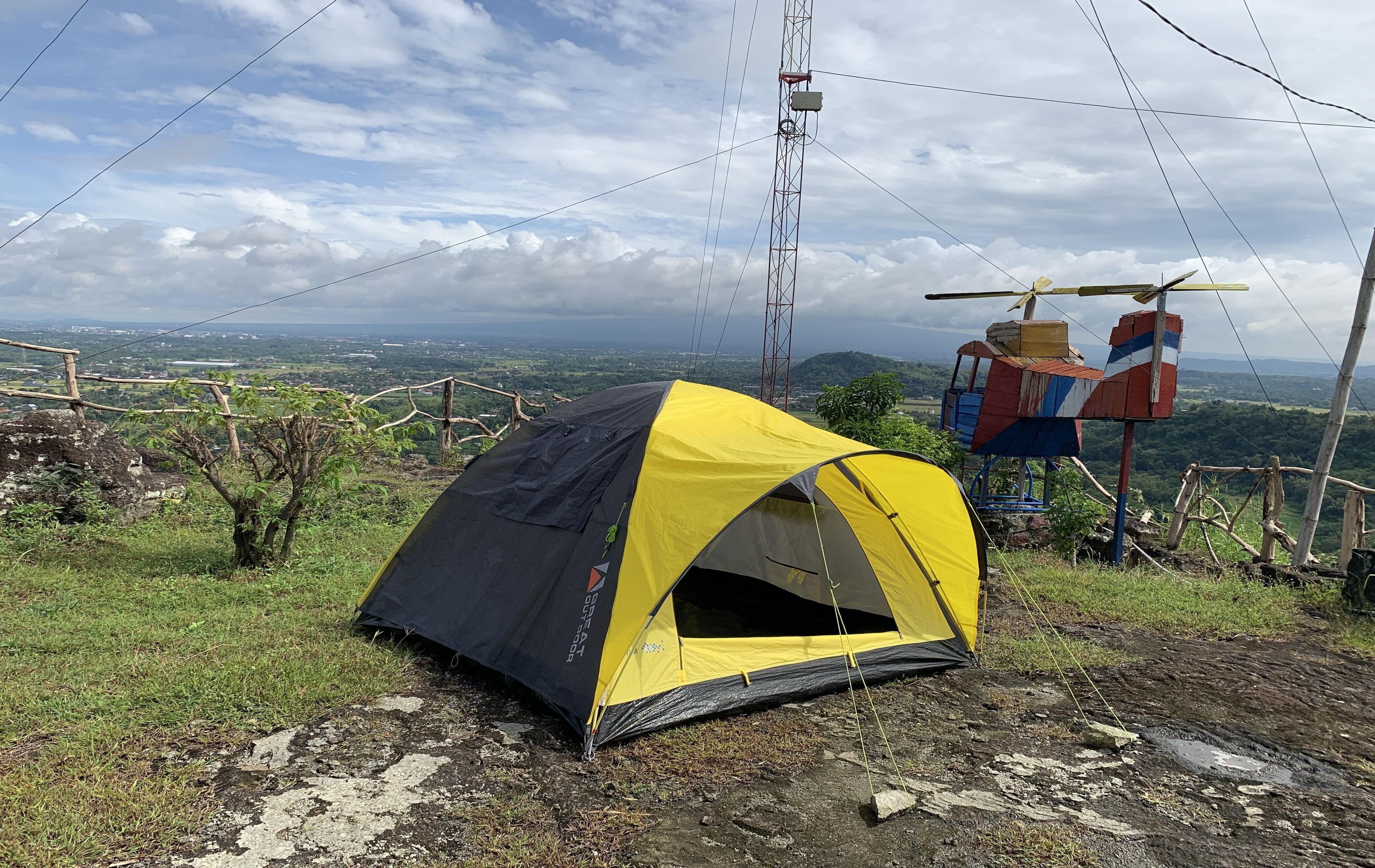 Una tenda gialla e nera montata su una collina erbosa, con vista sul paesaggio e una torre di linee elettriche sullo sfondo.