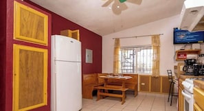 A compact kitchen with a refrigerator, wooden cabinets, a dining table, and a window with curtains.