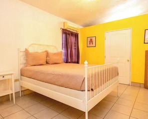 A single-bed room with a white metal frame, a beige bedspread, a small bedside table, a door, and a window with curtains.