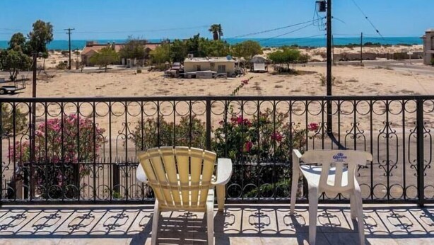 Ein Balkon mit zwei Stühlen und einem Tisch, der Blick auf einen Strand mit Gebäuden und einem klaren Himmel bietet.