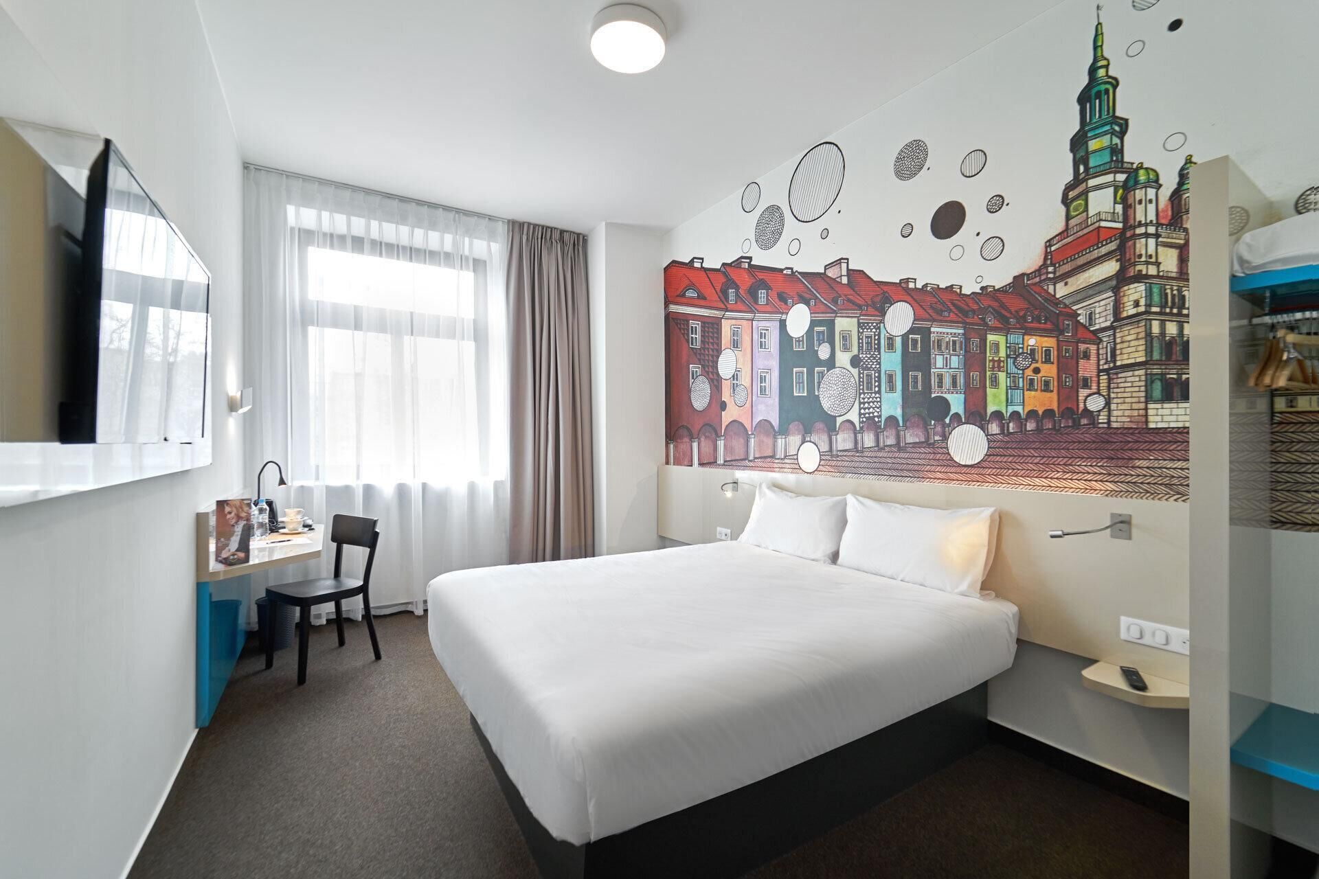 A hotel room with a large bed, a desk with a chair, a flat-screen TV, and a mural of a cityscape on the wall.