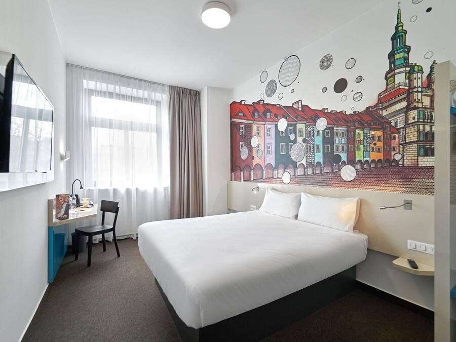 Une chambre d’hôtel avec un grand lit, un bureau avec une chaise, un téléviseur à écran plat et une fresque représentant un paysage urbain sur le mur.