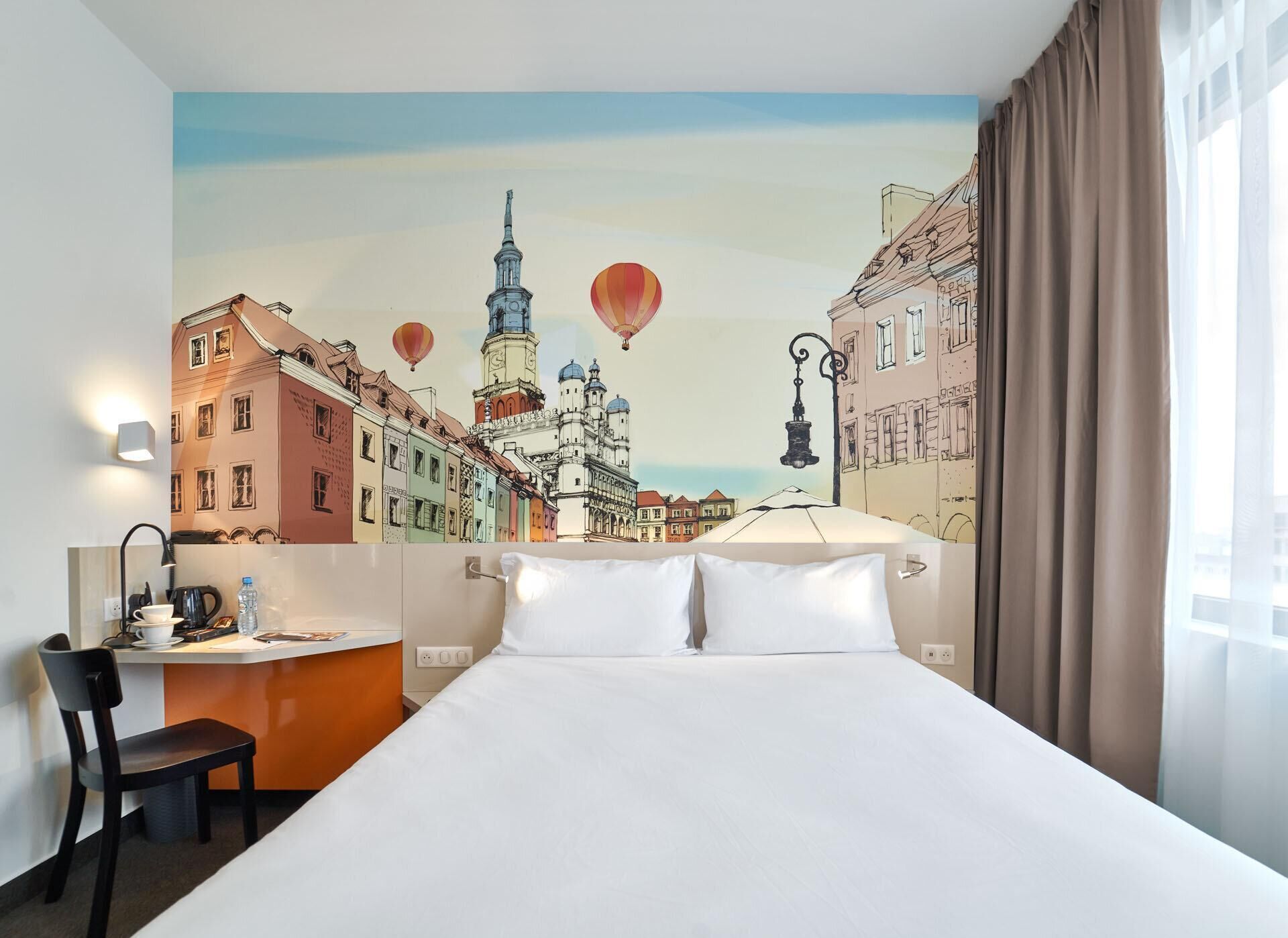 Ein Hotelzimmer mit einem Bett, einem Schreibtisch mit Stuhl, einer Lampe und einem Wandbild einer Stadtansicht mit Heißluftballons.
