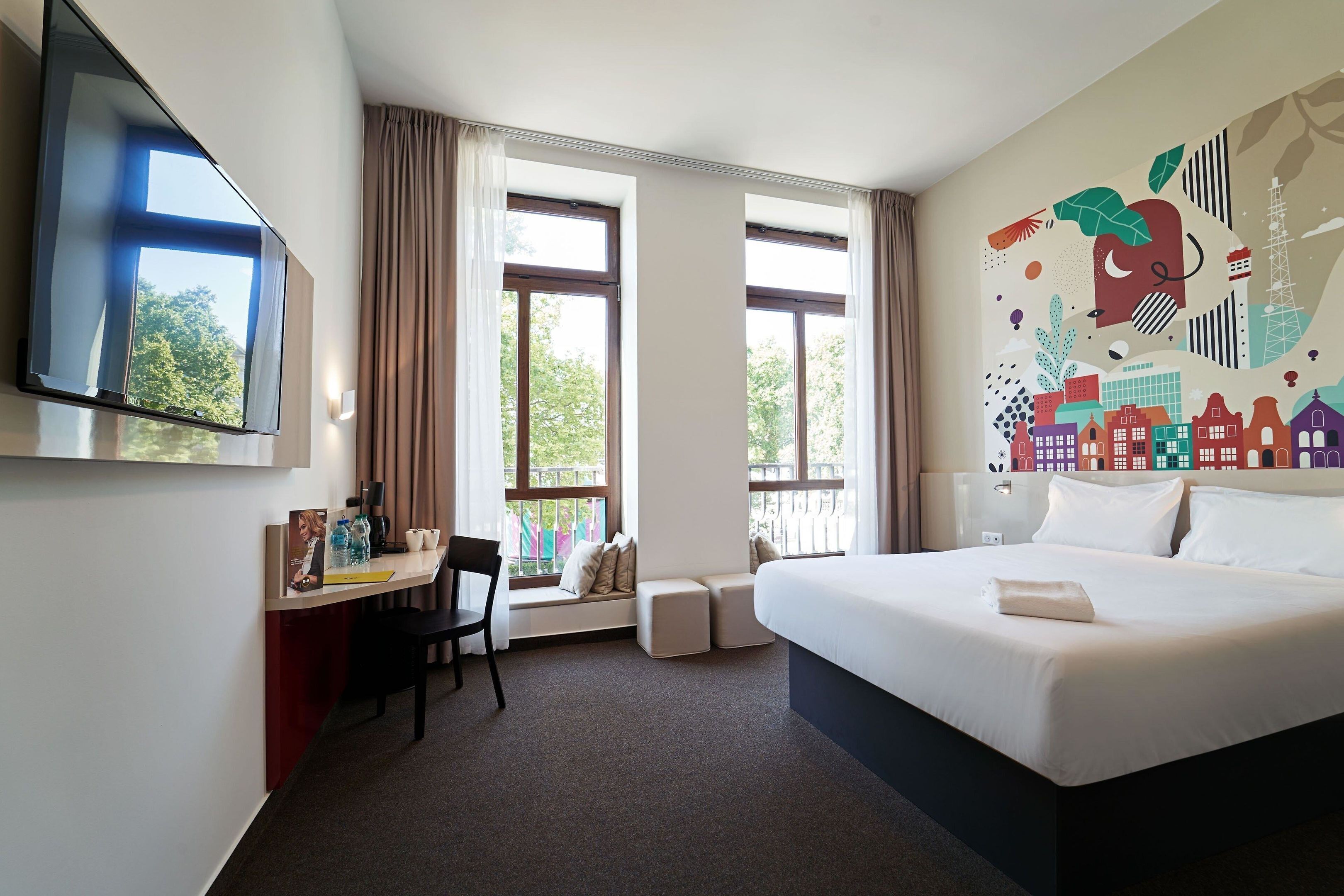 A hotel room with a bed, a desk with a chair, a TV, and large windows.
