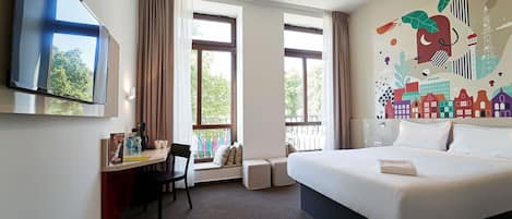 A hotel room with a bed, a desk with a chair, a TV, and large windows.