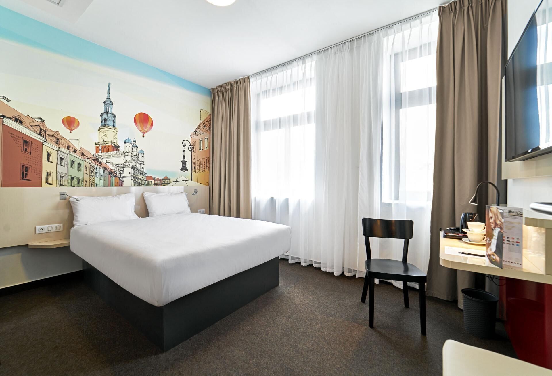 A hotel room with a bed, a desk with a chair, a TV, and a mural of a cityscape on the wall.