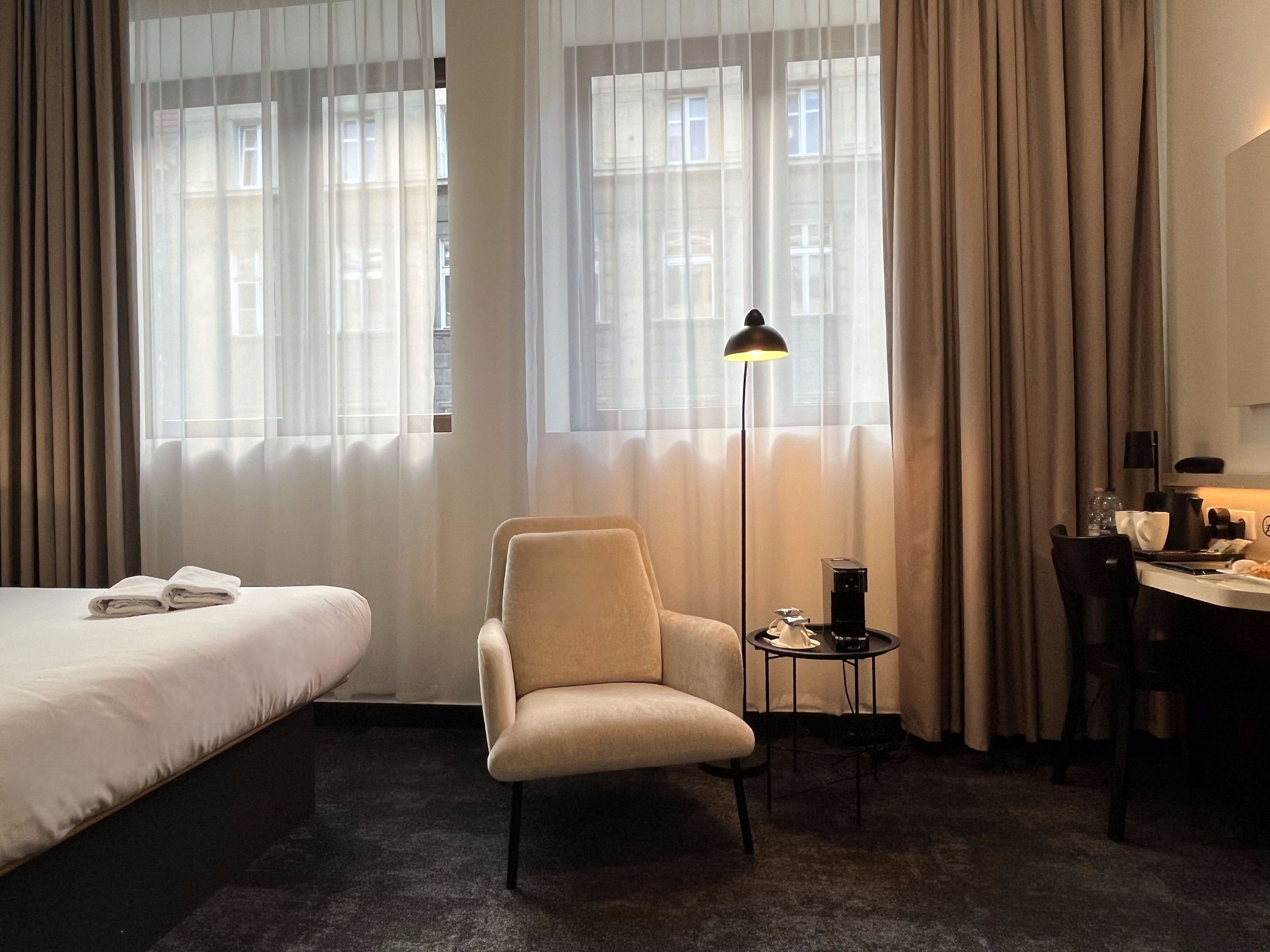 Ett hotellrum med en säng, en stol, ett skrivbord med en lampa och en kaffebryggare.