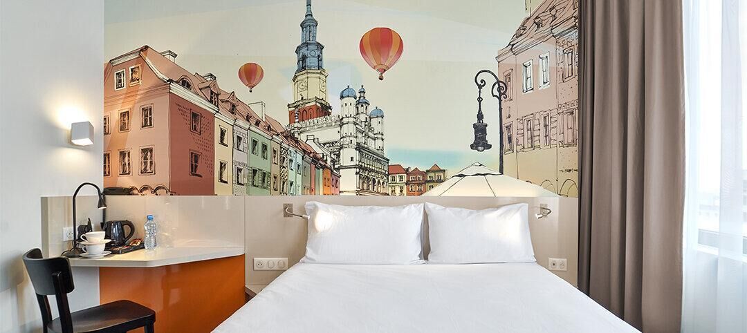 Habitación de hotel con cama, escritorio, silla y un mural en la pared que muestra un paisaje urbano con globos aerostáticos.