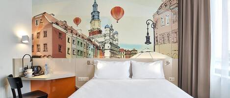 Habitación de hotel con cama, escritorio, silla y un mural en la pared que muestra un paisaje urbano con globos aerostáticos.