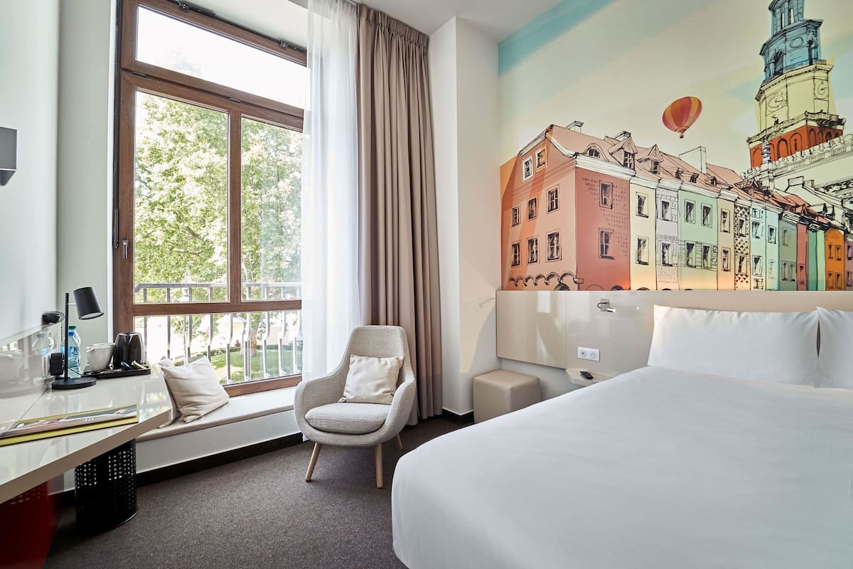 Hotelkamer met een groot raam, een bed, een stoel, een bureau met een lamp en een muurschildering van kleurrijke gebouwen.