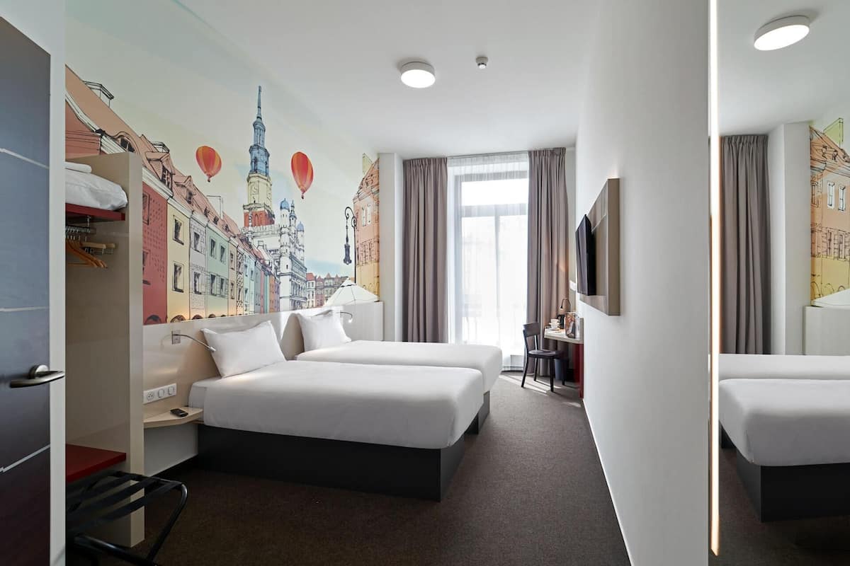 Een hotelkamer met twee bedden, een bureau, een stoel, een televisie en een muurschildering van een stadsgezicht.