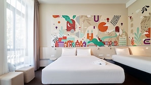 Une chambre d’hôtel avec un grand lit, deux tables de chevet, une fenêtre avec des rideaux légers et une fresque murale abstraite et colorée.