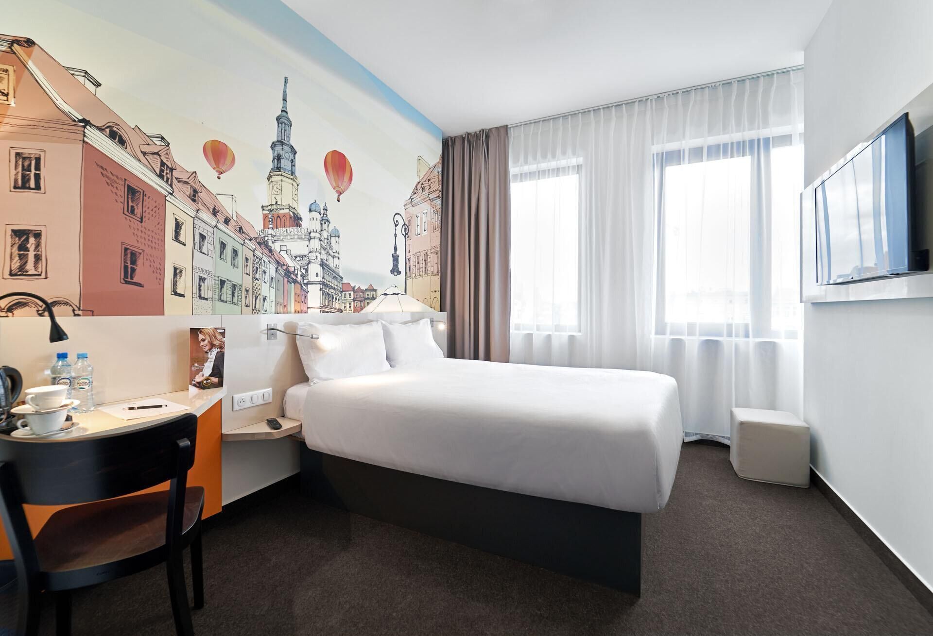 Ein Hotelzimmer mit Bett, Schreibtisch mit Stuhl, einem kleinen Tisch mit Tasse und Untertasse, einem Wand-Fernseher und einem Fenster mit Vorhängen.