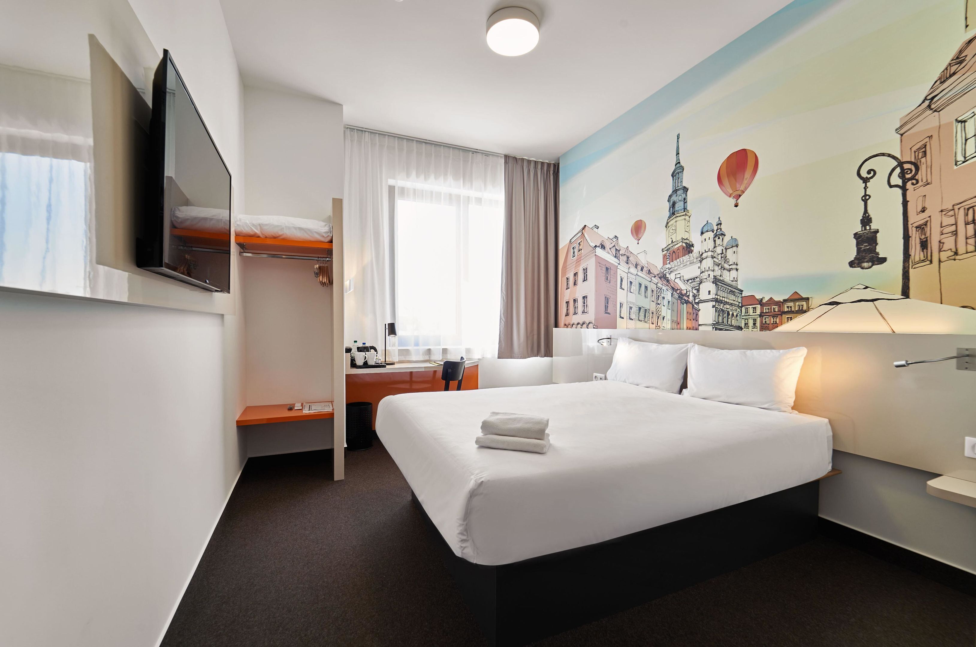 A hotel room with a large bed, a desk, a chair, a TV, and a mural of a cityscape.