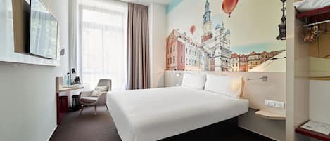 Quarto de hotel com uma cama grande, uma escrivaninha com cadeira, televisão na parede e um mural com vista urbana.