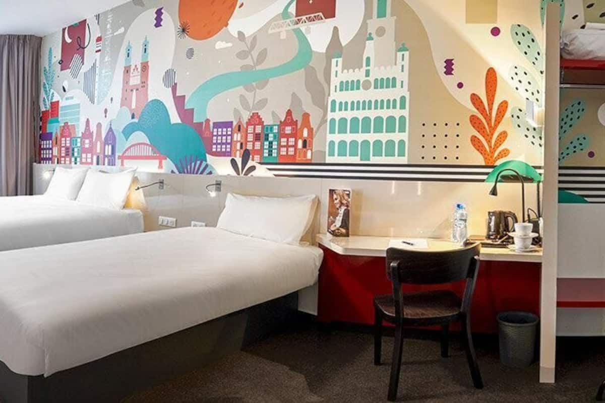 Een hotelkamer met twee bedden, een bureau, een stoel en een kleurrijke muurschildering.