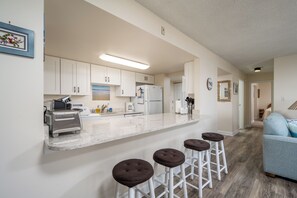 A modern kitchen with white cabinets, a marble countertop, and a built-in refrigerator. There are three stools at the counter and a light fixture above.