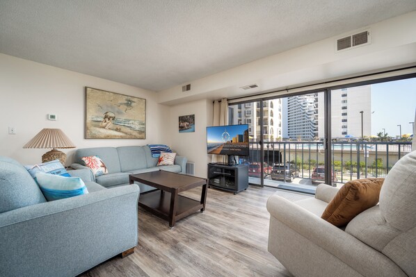 A modern living room with a sofa, a coffee table, and a TV. There is a large window with a view of buildings and a balcony.