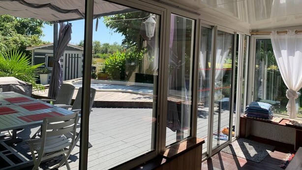 Une véranda dotée de grandes portes-fenêtres coulissantes, un espace patio avec une table et des chaises, et une vue sur un jardin avec une piscine et de la verdure.