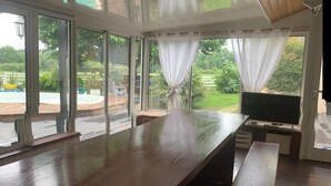 Une pièce spacieuse avec une grande table en bois, un téléviseur et de grandes fenêtres ornées de rideaux légers.