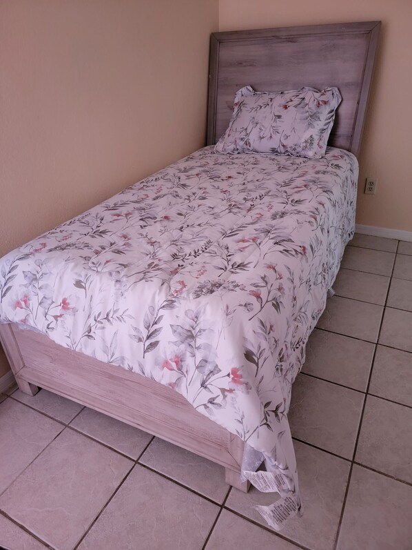 Un lit simple avec une couvre-lit à motifs floraux et une tête de lit en bois, dans une pièce dont le sol est recouvert de carreaux.