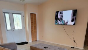 Ein Wohnzimmer mit einem beige-farbenen Ecksofa, einem kleinen Hocker, einem flachbildfernseher an der Wand und einer Tür mit Milchglasfenstern.