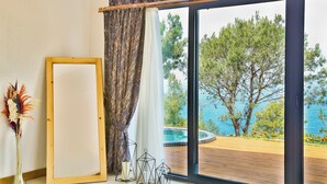 A room with a large window overlooking a pool and trees, a framed picture, and a vase with dried flowers. - Vista Del Amor Faralya (Fethiye)