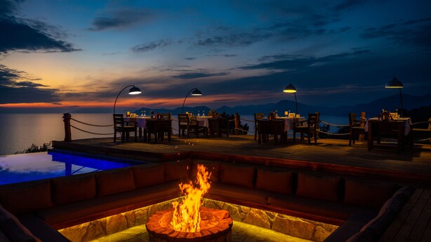 A poolside dining area with tables and chairs, a fire pit, and a view of the sea at sunset. - Vista Del Amor Faralya (Fethiye)