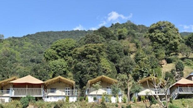 A row of thatched-roof cottages with balconies, set against a backdrop of dense forest and hills. - Camp Cedar (Baijnath)