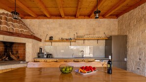 A rustic kitchen with a stone wall, wooden beams, a brick fireplace, and a wooden table with fruit and a wine bottle. - Villa Petar ZadarVillas (Donji Kašić)