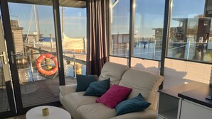 Una sala de estar moderna con un sofá color beige, una mesita redonda y vista a un muelle con barcos.