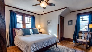 Quarto com cama, ventoinha no teto, mesa de cabeceira, cadeira e janela com cortinas.