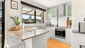 A modern kitchen with white cabinets, granite countertops, a built-in oven, and a window overlooking a green landscape. - Back Beach Beauty in Smiths Beach (Smiths Beach)