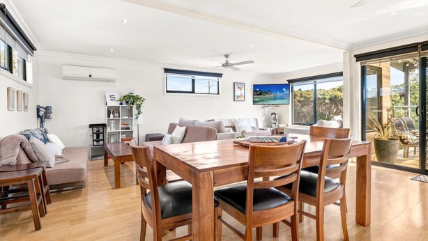 A spacious living area with a wooden dining table, comfortable seating, a ceiling fan, and large windows offering a view of the outdoors. - Back Beach Beauty in Smiths Beach (Smiths Beach)