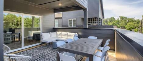 Relax on the outdoor deck with a wooden table, comfortable seating, and a sofa, surrounded by a patterned rug and a glass door leading to a covered patio.