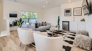 Uma sala moderna com sofá, duas poltronas brancas, mesa de centro, televisão, ventilador de teto, área de cozinha com geladeira e uma janela grande.