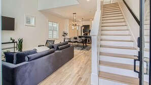 A modern living room with a grey sofa, wooden staircase, and a dining area in the background. - 17- Home w/ Rooftop - The Music City Collection (Nashville)