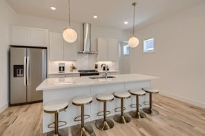 Una cocina moderna con una isla central, electrodomésticos de acero inoxidable, iluminación colgante y taburetes de bar.