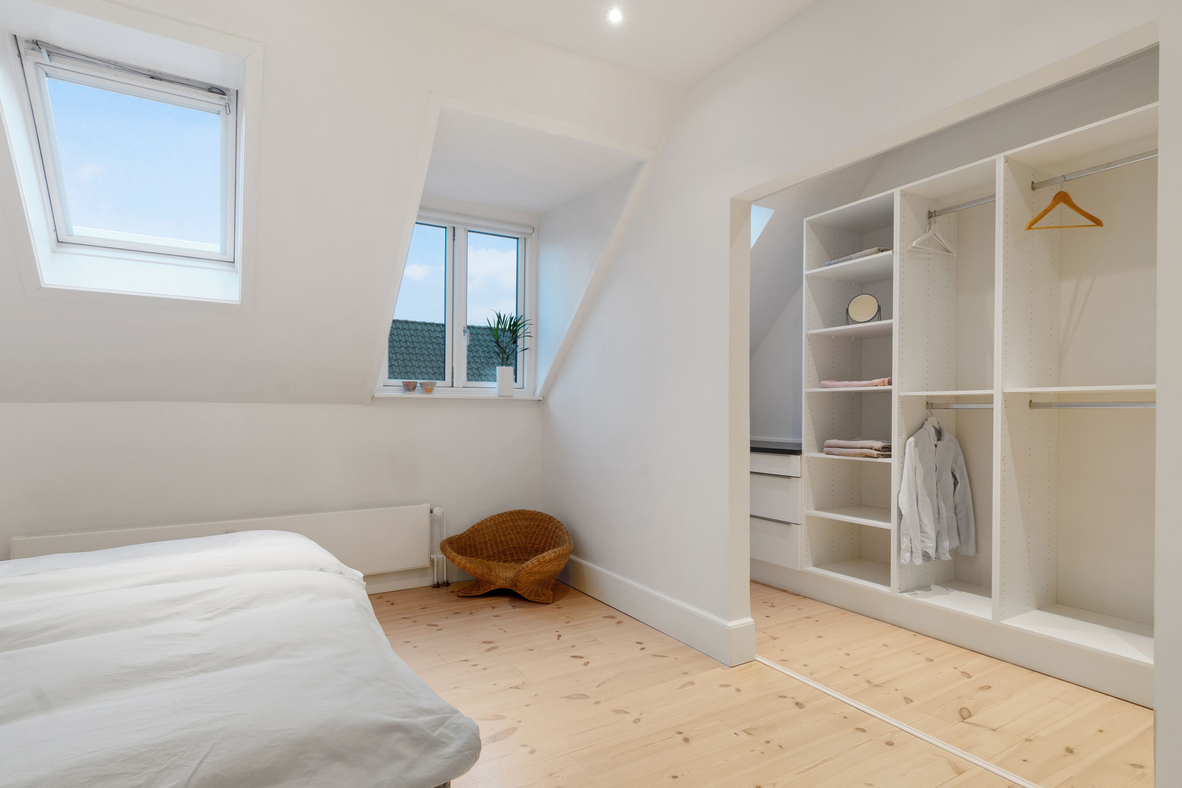 A bedroom with a bed, a window, a wooden floor, a wicker chair, and a walk-in closet with shelves and hangers.