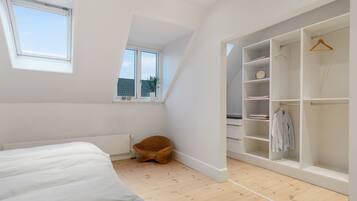 A bedroom with a bed, a window, a wooden floor, a wicker chair, and a walk-in closet with shelves and hangers.