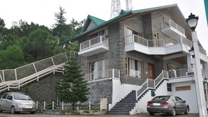 A two-story building with a stone facade, a green roof, and a balcony on the upper floor. There are two cars parked in front. - Echor The Rustic Retreat Kasauli (Kasauli)
