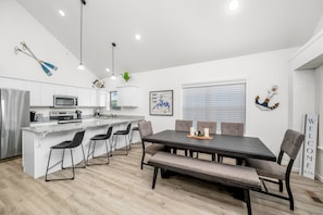 Une cuisine moderne avec des meubles blancs, un îlot central doté de tabourets de bar, une table à manger avec des chaises et une pancarte indiquant « BIENVENUE ».