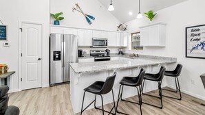 Een moderne keuken met witte kasten, roestvrijstalen apparatuur, een granieten aanrechtblad en zwarte barstoelen.