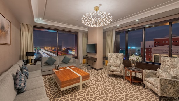 Habitación de hotel moderna con un ventanal grande que ofrece vistas a la ciudad, un sofá, una mesa de centro y una alfombra estampada.
