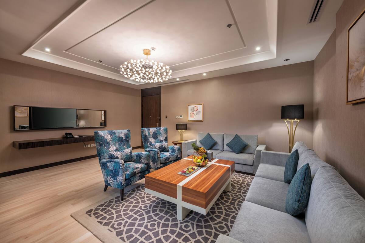 Ein modernes Wohnzimmer mit einem Holztisch, blauen Sesseln mit Muster, einem grauen Sofa, einem großen Flachbildfernseher und einer Deckenleuchte.