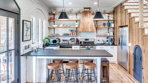 A modern kitchen with a large island, bar stools, wooden shelves, and a built-in stove. - Carlton Landing-The Coral Cottage (Carlton Landing)