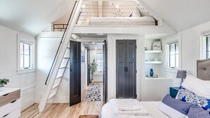 A loft bedroom with a wooden floor, a bed with white bedding, a blue and white patterned pillow, a nightstand, and a staircase leading to an upper loft area. - Carlton Landing-The Coral Cottage (Carlton Landing)