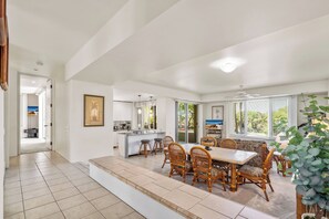A spacious dining area with a large table and wicker chairs, a kitchen with a white countertop, and a view of the outdoors through large windows. - Palms at Wailea #1605 Tropical Garden View Ground Floor, Wifi, Shared Hot Tub (Wailea)